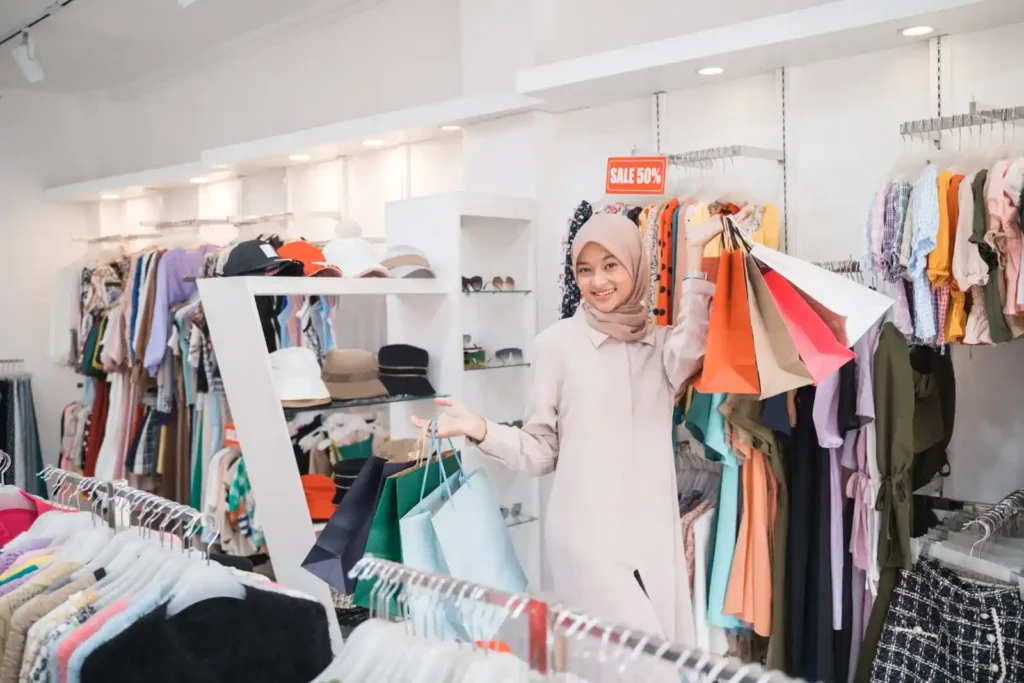 Wanita berhijab tersenyum memegang beberapa tas belanja di toko pakaian dengan tulisan "SALE 50%