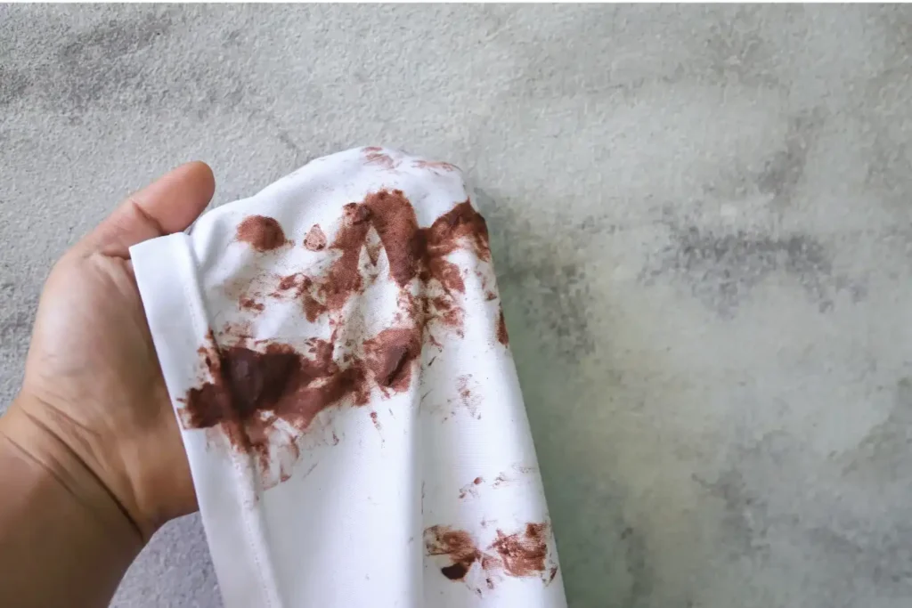 Tangan memegang sepotong kain putih dengan noda cokelat yang terlihat membandel, dengan latar belakang dinding bertekstur.