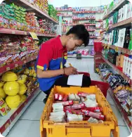 Karyawan Toko Yan sedang mencatat stok barang di lorong toko dengan keranjang penuh produk.